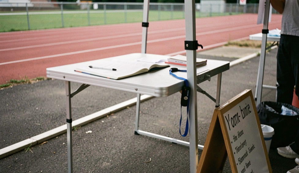 A folding table under a canopy at the edge of a track, an open notebook, a pen on a lanyard and a sign-in board leaning against the table leg