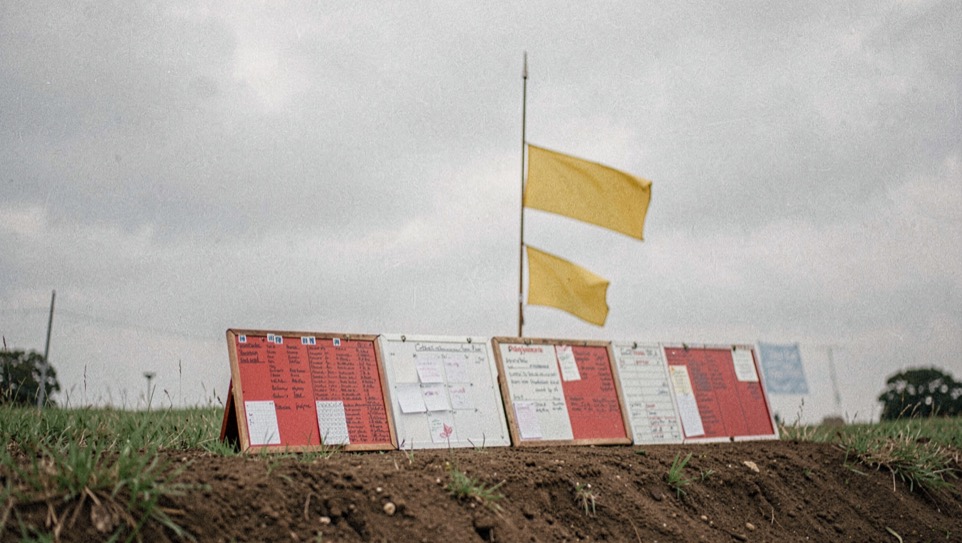 A row of red and white marker boards along the top of a berm, one leaning, with a yellow flag tied to a thin pole against an overcast sky